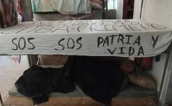 Una litera de metal con una sábana blanca colgada de la parte superior, mostrando mensajes escritos en negro como "SOS SOS Patria y Vida"